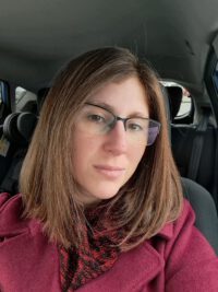 Frontalbild von Katrin Biasi, im Auto sitzend. Etwas über Schulterlänge braune Haare, roter Wintermantel.